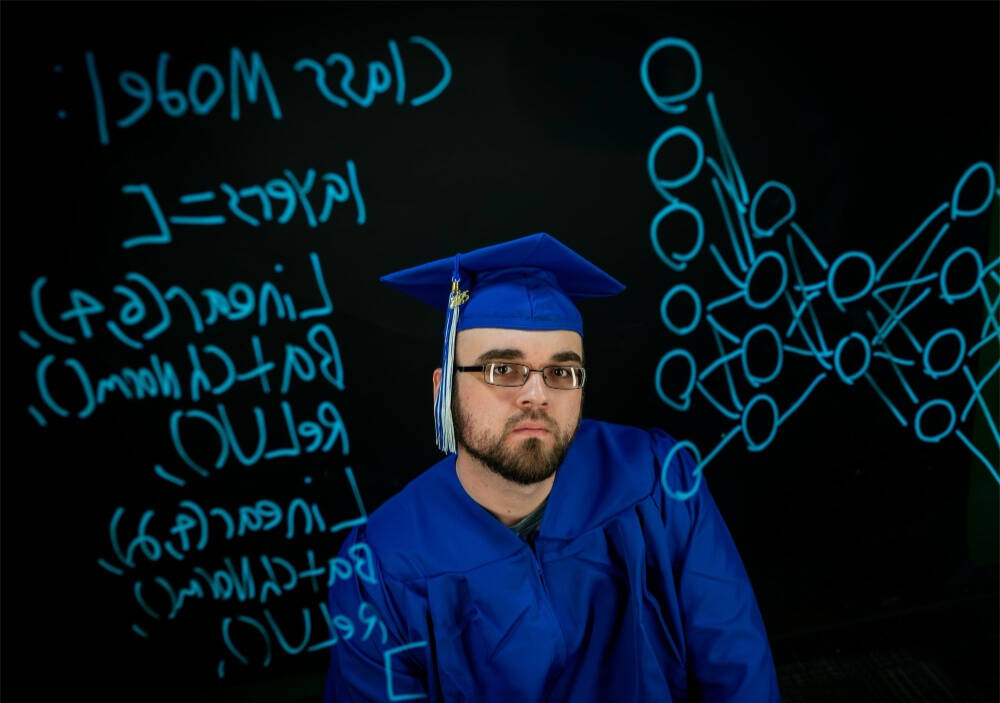 Tony Boos is wearing a blue graduation cap and gown, standing in front of a blackboard with neon blue scribbles and diagrams